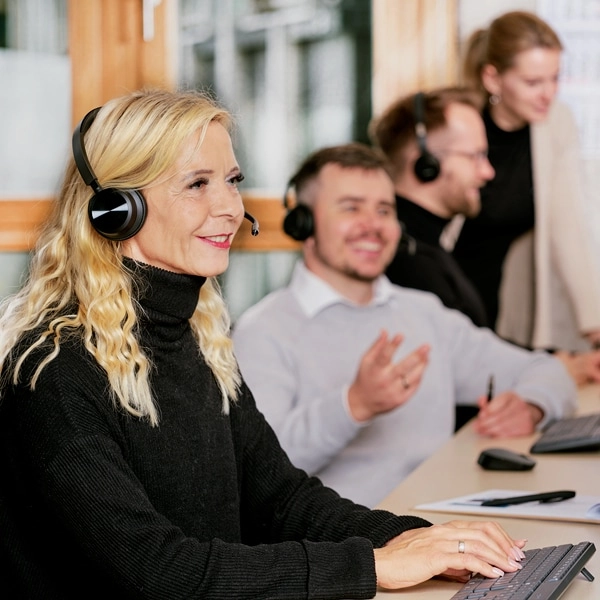 Eine Blonde Frau aus dem Customer Management Team führt offenbar ein Kundengespräch mit Headset. Im Hintergrund sind drei weitere Personen aktiv in Gesprächen involviert