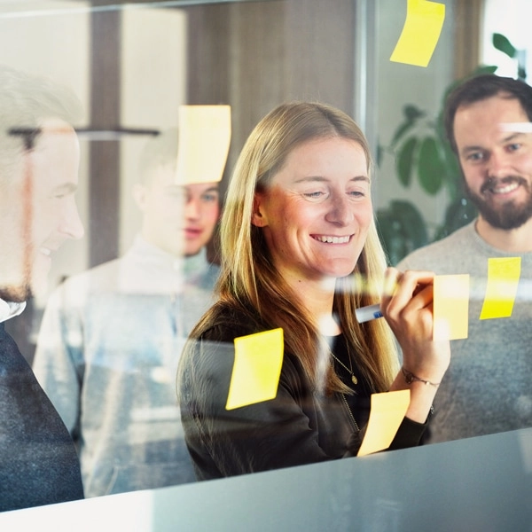 im Vordergrund stehen ein Mann und eine Frau aus dem Strategic Projects & Business Developtment Team an einer Glaswand. Sie führen dabei ein Gespräch und schreiben etwas auf Post-it Zetteln.